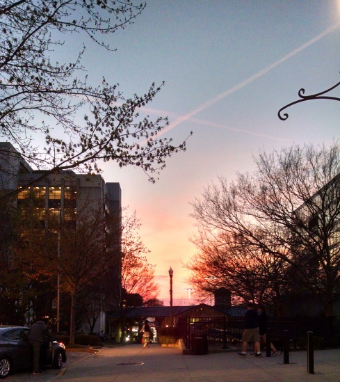 The sun sets behind low buildings and a sidewalk. On the left rises a taller building and a few budding trees. The sky is orange fading to blue and purple, and two airplane trails make a gold "x" in the sky.