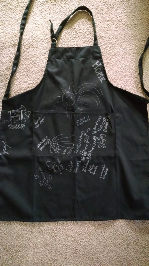 A black apron is spread out on a carpt with various affirmations written in silver ink. There is a picture of a cow with the word "vegan" and a heart; "nurturer"; "abundant generosity"; "open... a heart wide open" with a drawing of a heart; "WWE champions pretty" with a picture of a face with long hair; "me and Adrian rock around in the car boom yeah"; "sweetness"; "wholeness"; "silly Quaker"; "ardent hospitality"; "conscientious industrious"; a picture of a shooting star; "home"