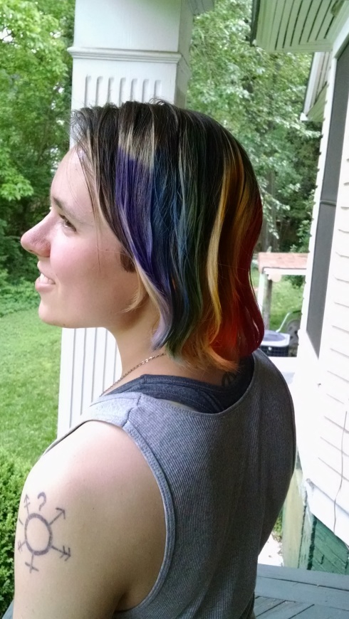Adrian, author of this blog, stands outdoors on a porch and faces to the left, displaying shoulder-length hair dyed rainbow stripes.